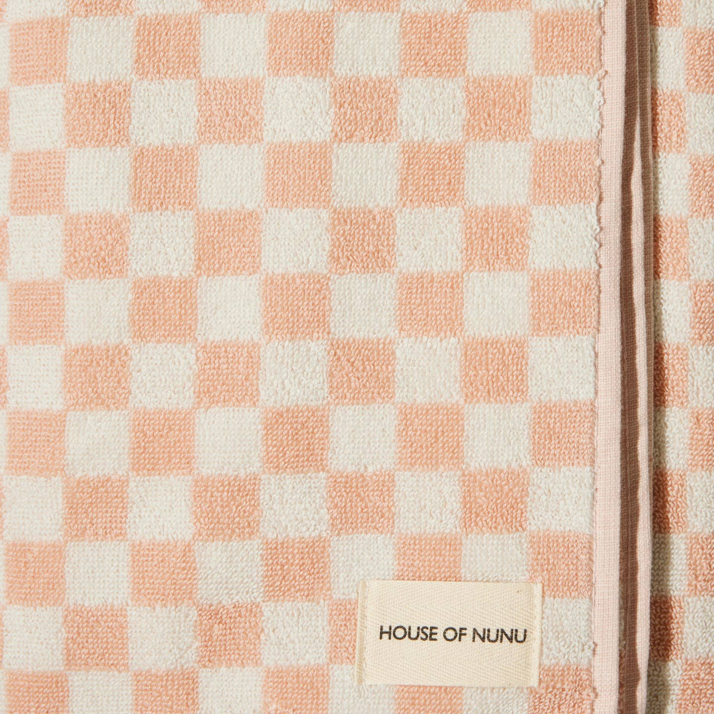 A close up of a bath towel in Pink Check by House of Nunu. This towel is white with soft peach checks and folded into a perfect rectangle. 