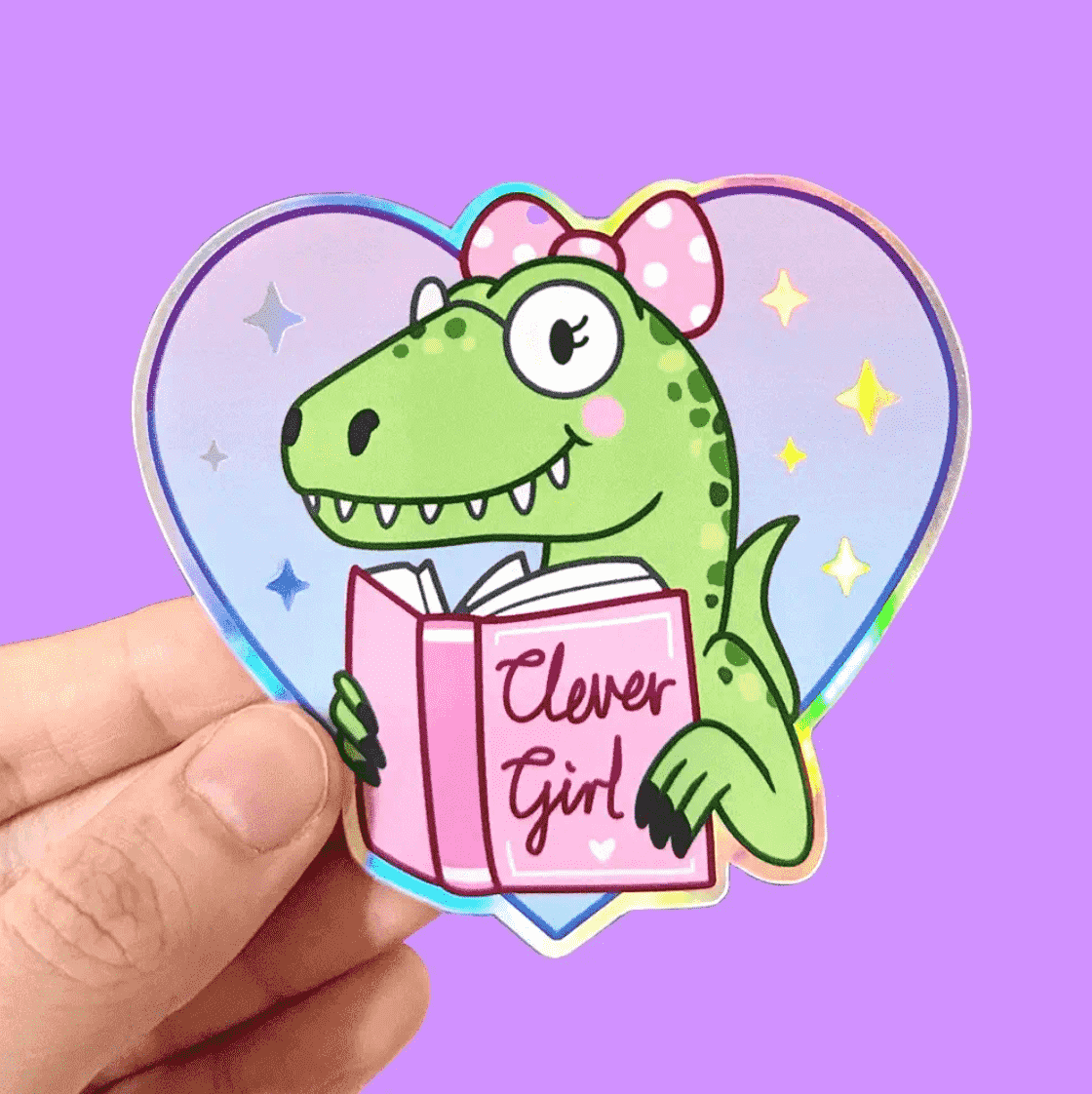 A sticker of a super cute, green dinosaur with a pink bow in their hair. They have glasses on and pink cheeks with a pink book that reads "Clever Girl" on the cover. Outside the dino is a purple heart with gold stars inside. 