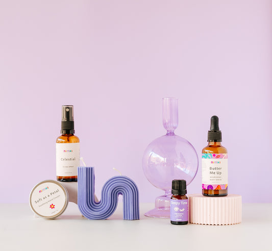 A collection of Nimki Aromatherapy and skincare products on a lilac background with pastel plinths elevating two of the products. 