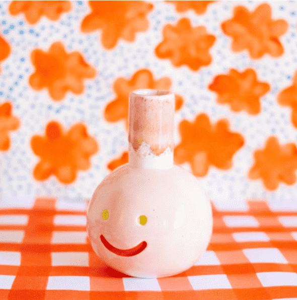 Shuh Lee's Suzette Vase: A super cute vase with a pastel pink exterior, yellow eyes and a red happy mouth. The background is a pattern adorned with orange daisies and blue speckles in between. 