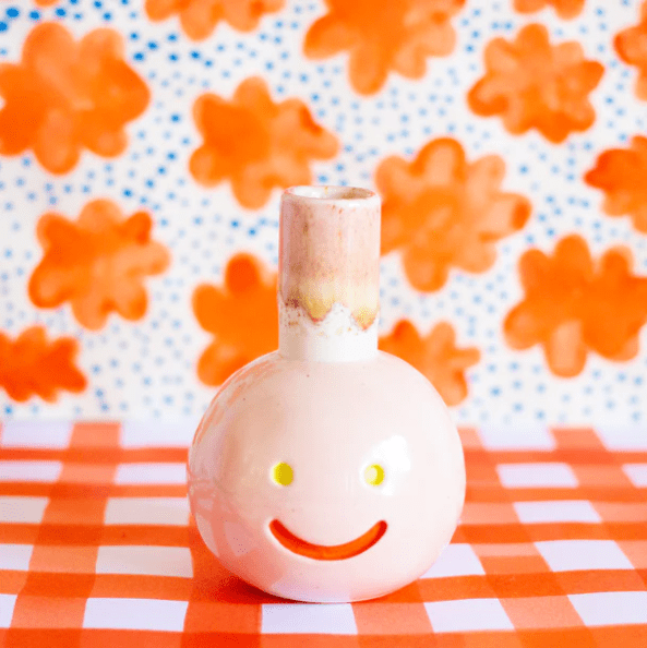 Shuh Lee's Suzette Vase: A super cute vase with a pastel pink exterior, yellow eyes and a red happy mouth. The background is a pattern adorned with orange daisies and blue speckles in between. 