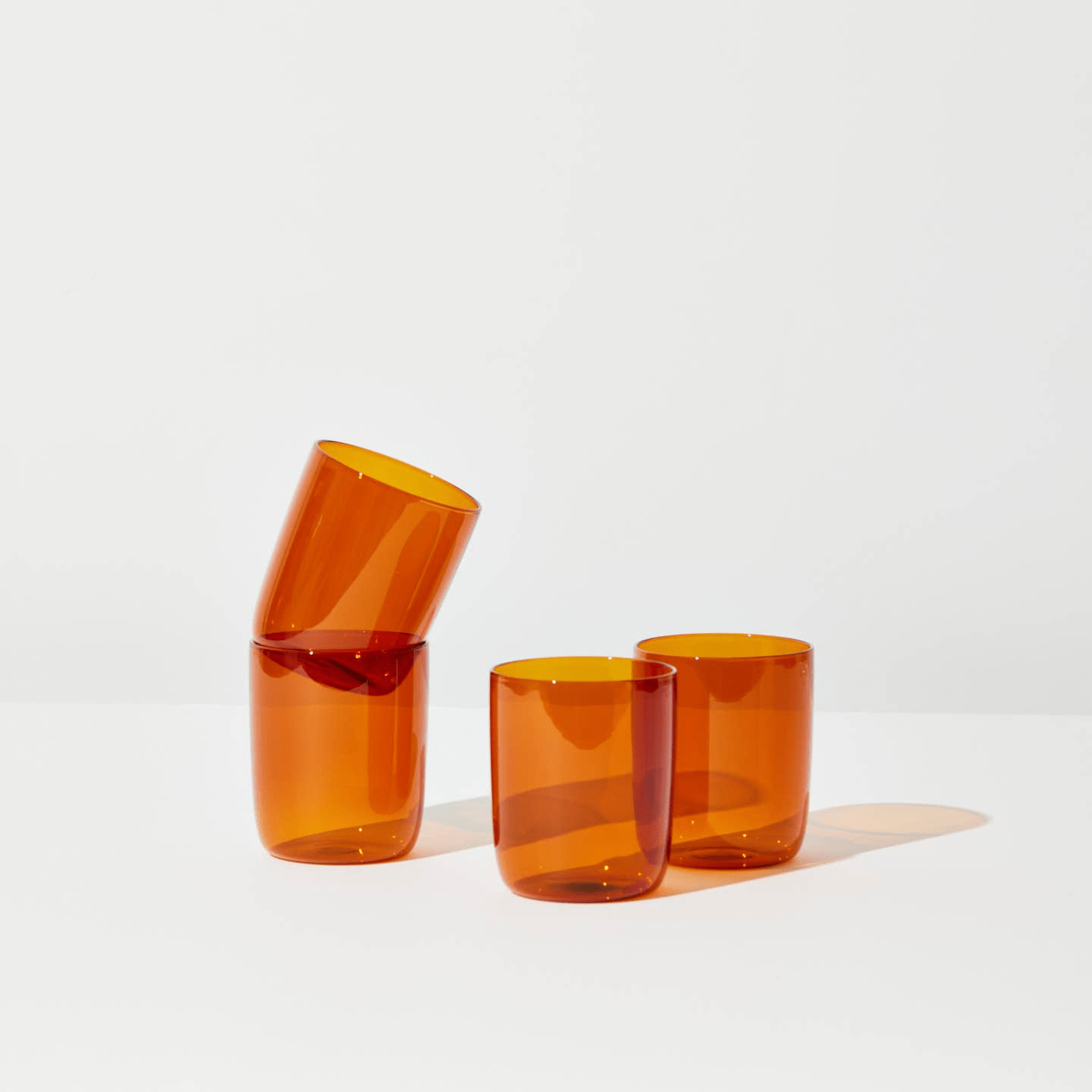 House of Nunu Amber Glass Set: Four amber coloured drinking glasses stacked on top of their brown box in front of a white backdrop. 