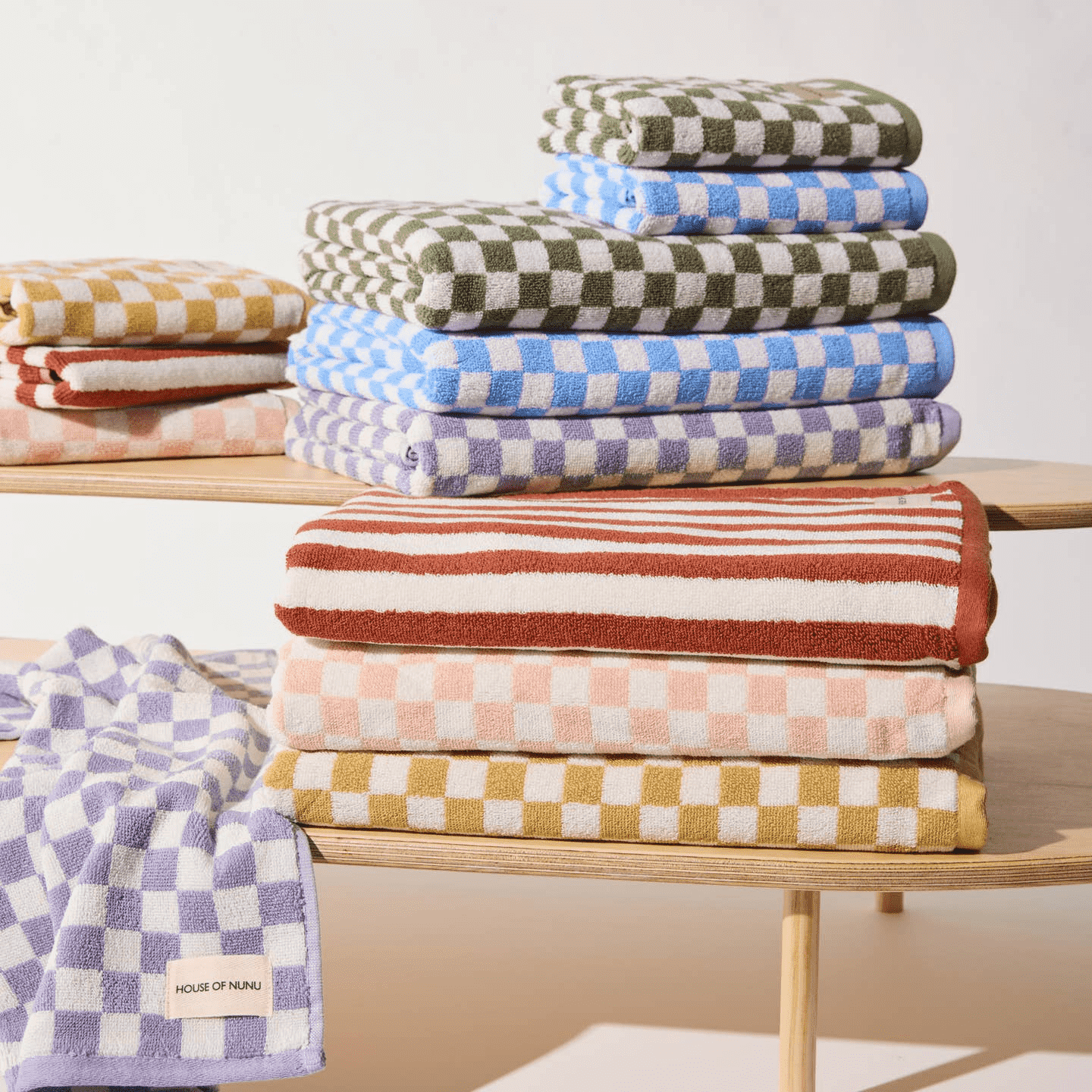 A colourful collection of House of Nunu's bath towels folded neatly on top of one another. It includes pastel coloured check prints and an orange stripe. 