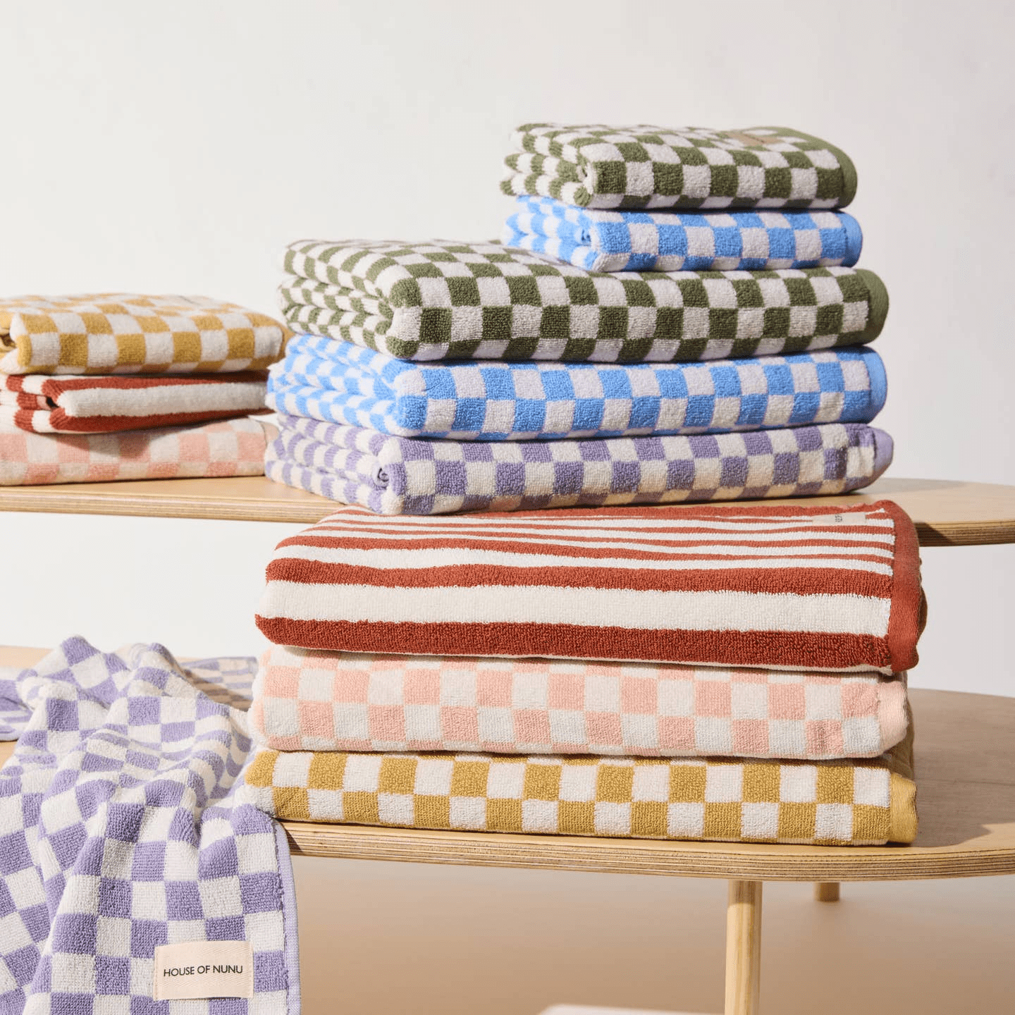 A super cute collection of House of Nunu bath towels and hand towels stacked on one another. Featuring pastel lilac, blue, forest green, peach, yellow checks and a red stripe print. 