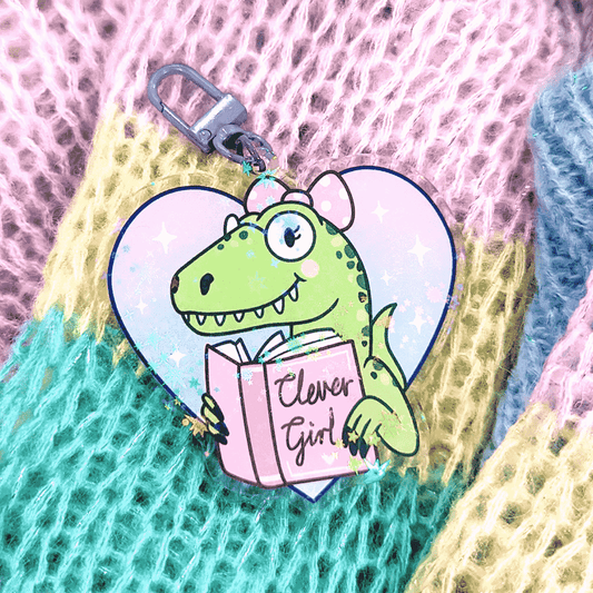 A cute keychain by Fox & Cactus of a happy green dinosaur. They have round glasses on, pink cheeks, a toothy grin and a pink bow in their hair. They have a book that reads "Clever Girl" 