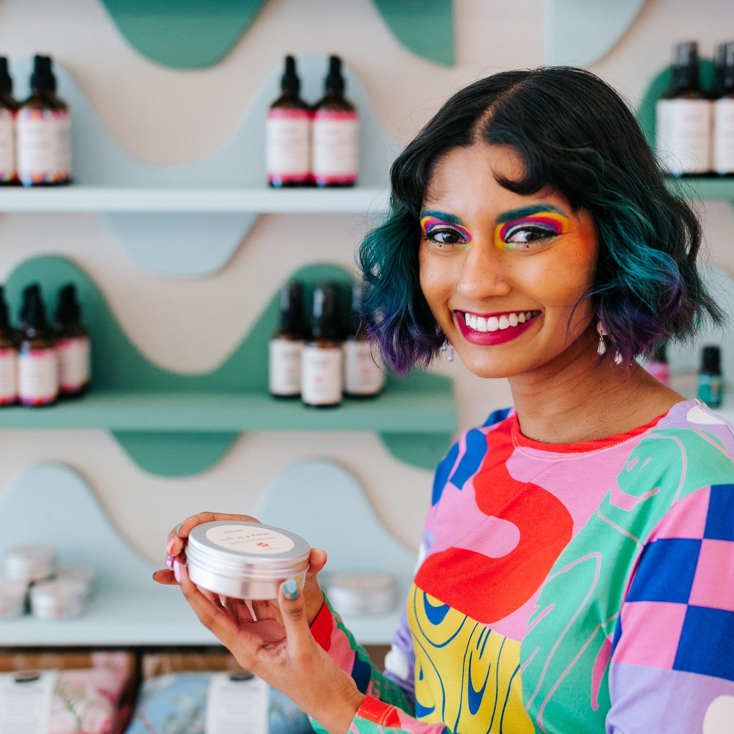 Nimki model Eishi holding the Sacred Wild Body Whip in a large circular tin, standing in front of Nimki's aqua blue and forest green wiggle shelves.