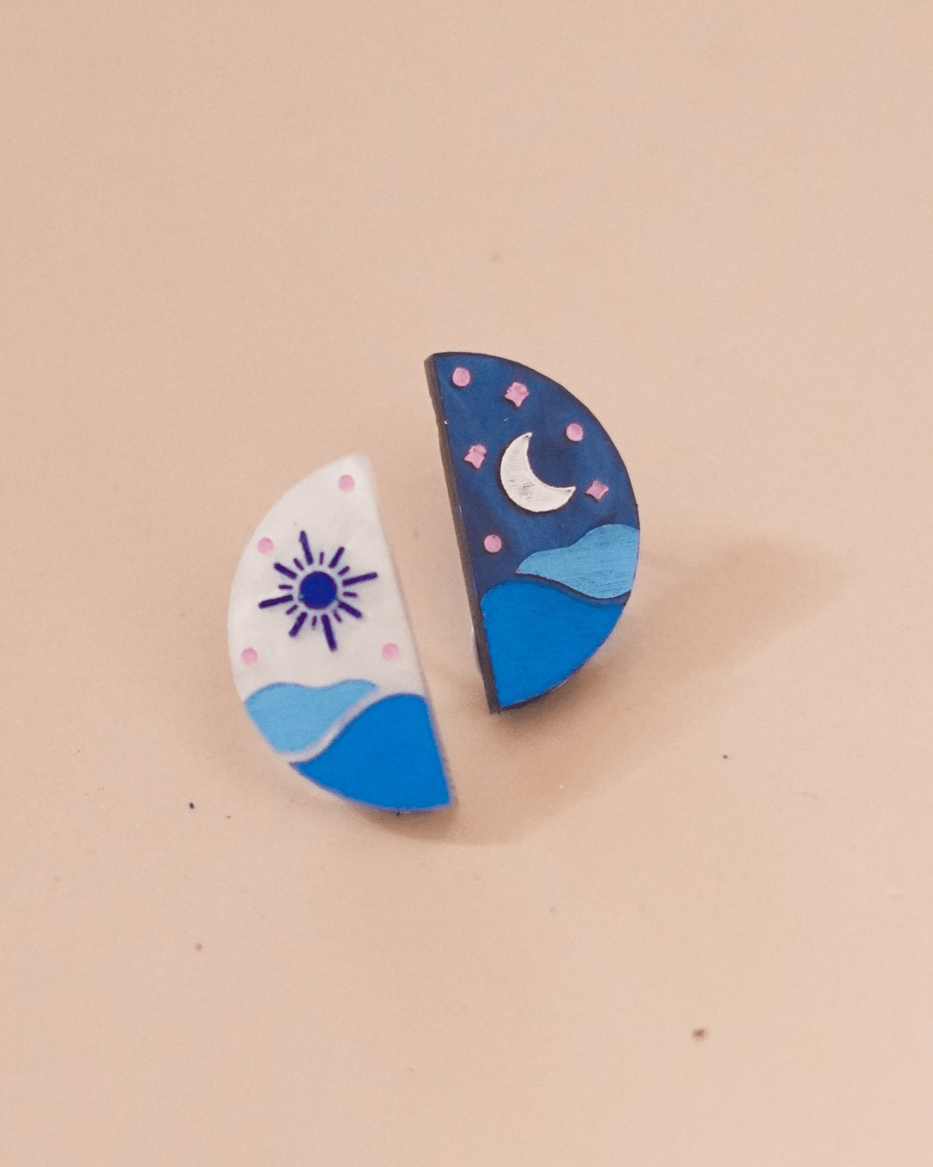 Flowatures dusk stud earrings. Each earring is one half of the other in a perfect half circle. Half is a sun with a white backdrop and blue waves while the other is a blue sky with a silver moon and blue sea. 