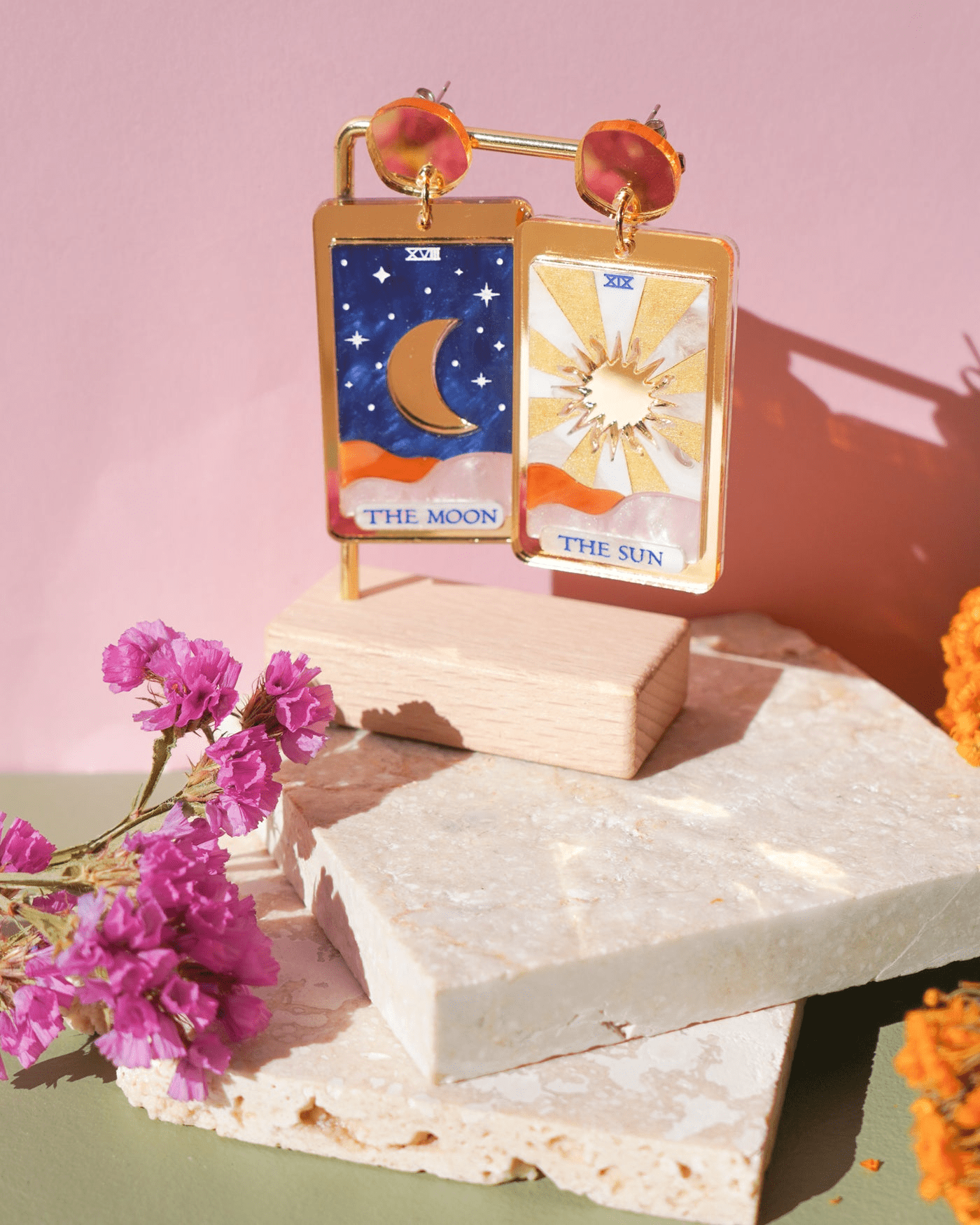 Flowature earrings moon and sun together with a pink backdrop behind them and stone props with flowers underneath. 