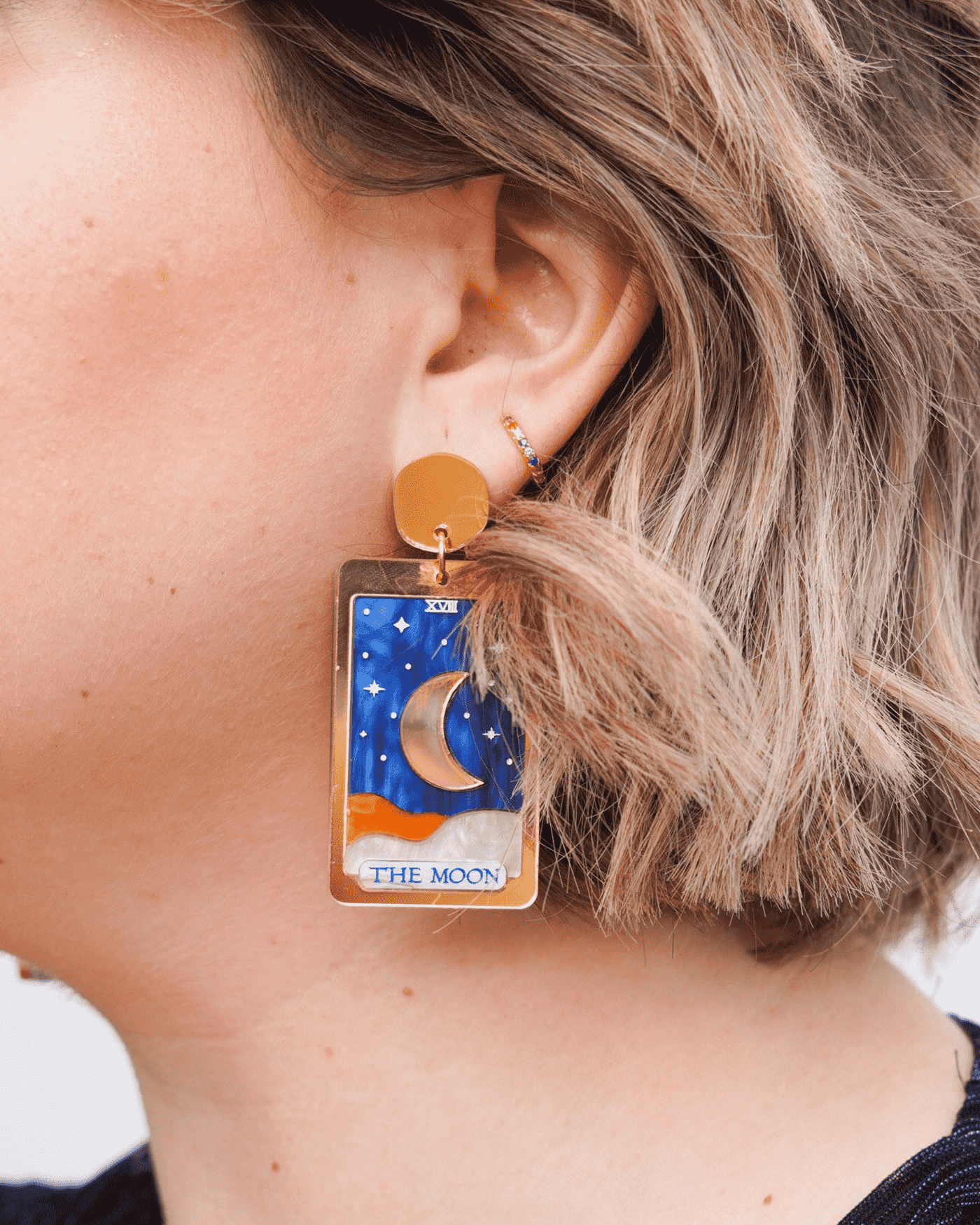 Flowature earring: a gold rectangular frame with a central gold slit moon surrounded by a blue sky with silver stars. Underneath it is a sea of orange and silver waves joining together and the words "The Moon" at the bottom. 