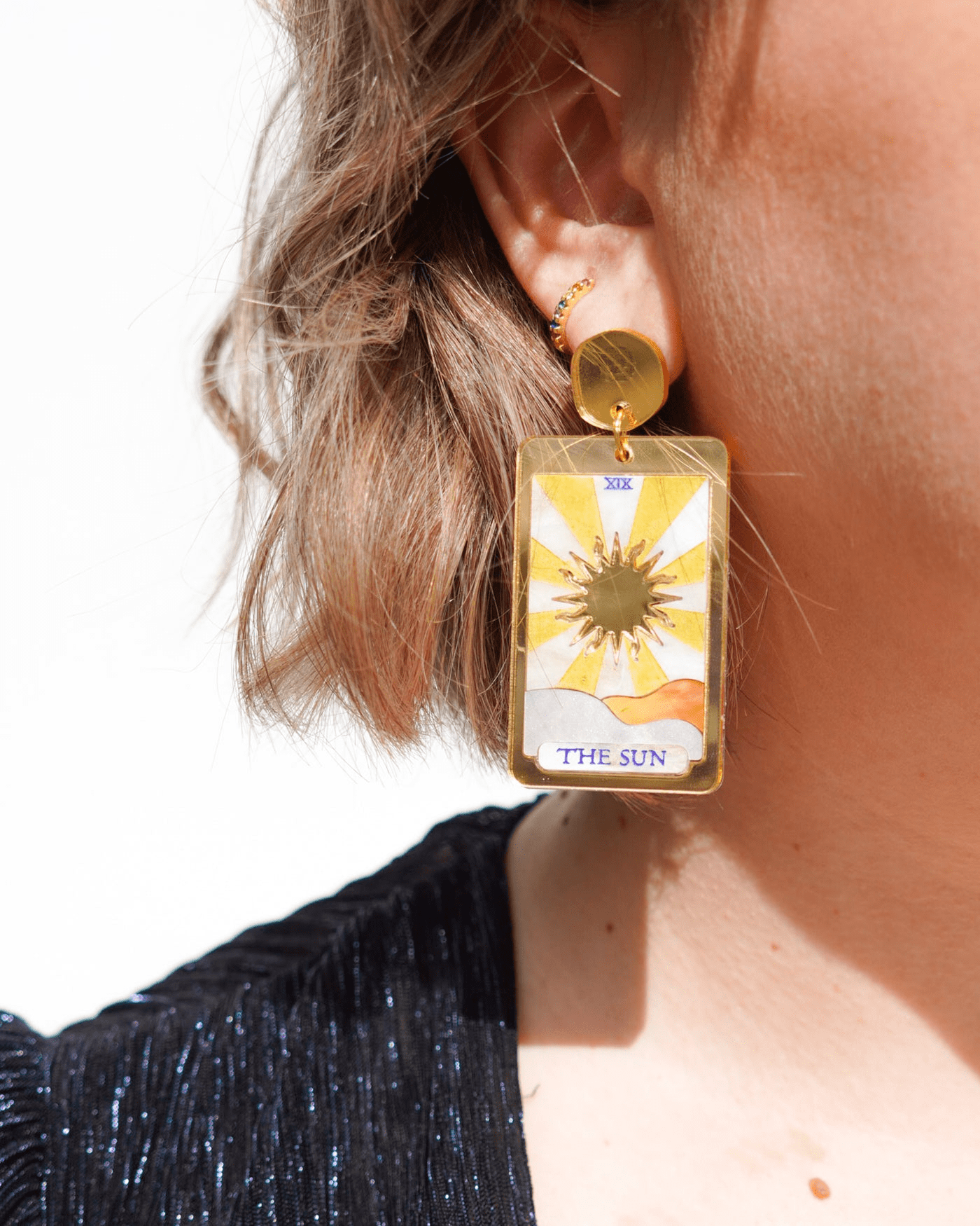 Flowature earring: a gold rectangular frame with a central gold sun surrounded by a yellow and white rays. Underneath it is a sea of orange and silver waves joining together and the words "The Sun" at the bottom. 