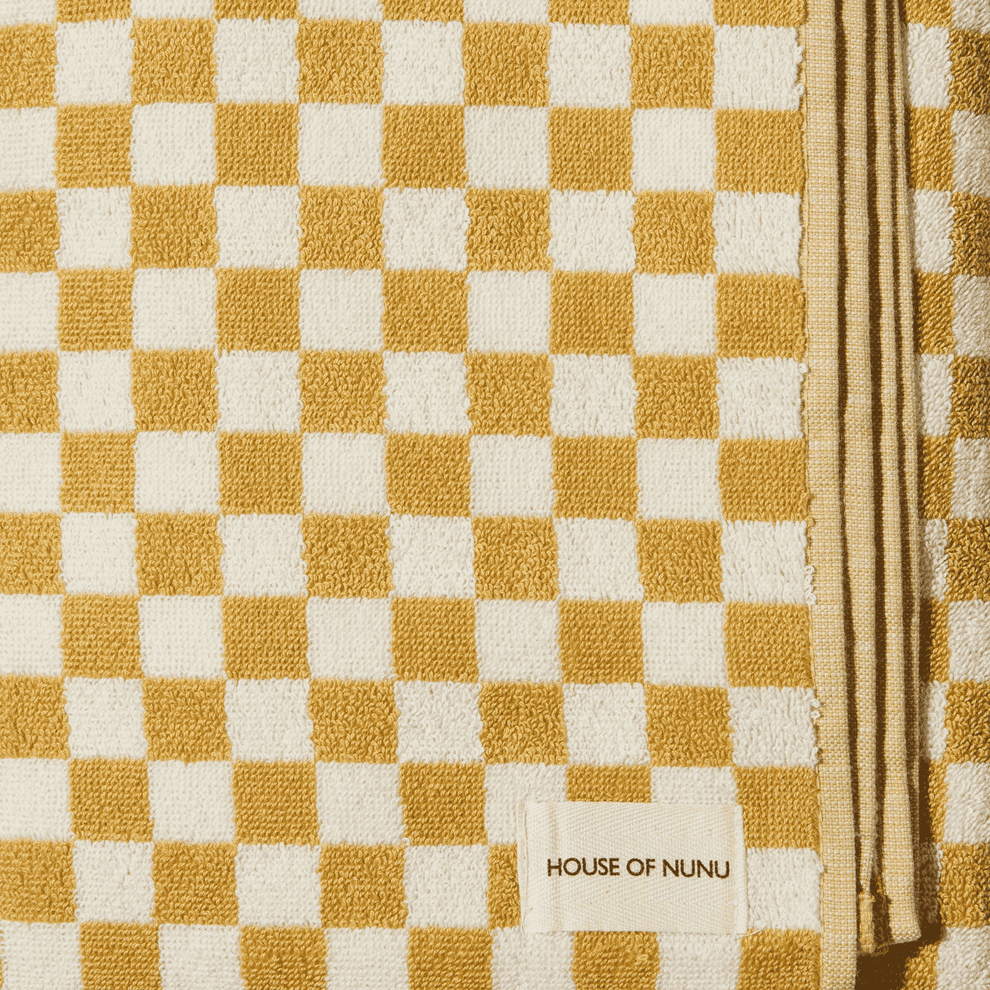 A close up shot of a House of Nunu bath towel: Folded into a perfect rectangle this towel features a white background with mustard yellow checks. 