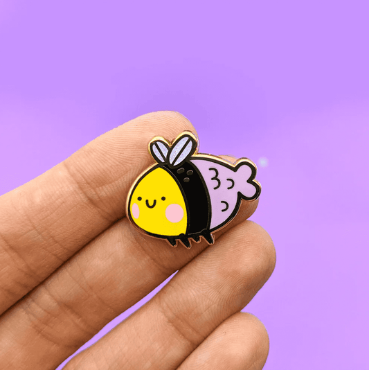 A pin with a bee shaped body and face, a pink mermaid tail and tiny insect like wings. It has a happy face and rosy cheeks. 