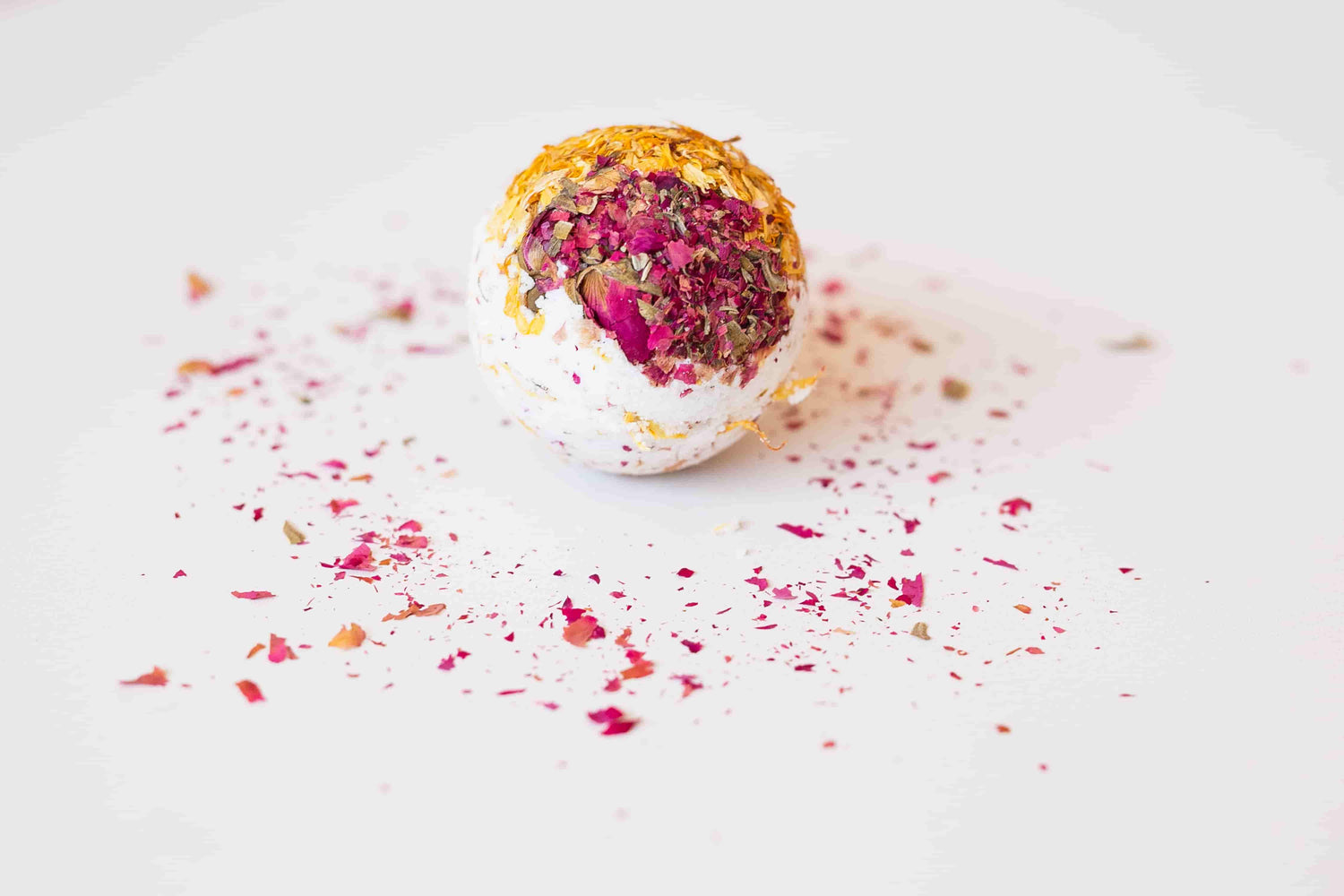 Nimki's natural aromatherapy bliss bath bomb sits on a cream tablecloth. The white bath bomb is topped with vibrant pink rose buds and petals on one half, and golden yellow calendula on the other. Scattered rose petals surround it for a touch of elegance.