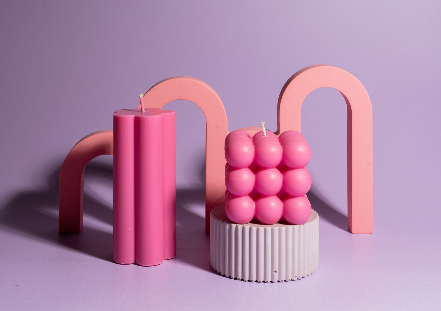 A pastel lilac backdrop featuring a pink arch prop in the background. On a lilac plinth sits a vibrant hot pink bubble-shaped candle alongside a hot pink flower-shaped pillar candle.