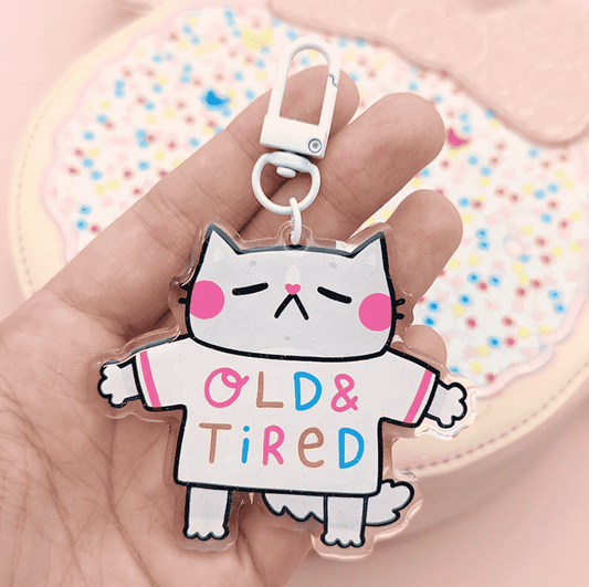 A super cute keychain by Fox & Cactus: a sleeping cat with pink cheeks and a heart note with a tee shirt that reads "Old & Tired" on the front and "Go Away" on the back. 