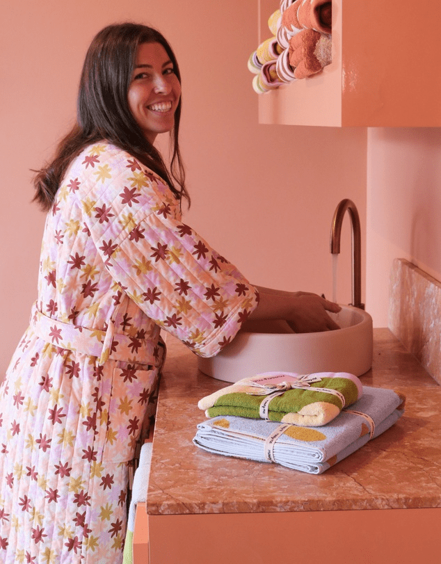 A woman with a big cheesy smile and long brown hair stands at a peach coloured basin washing their hands wearing the peach robe with pride. 