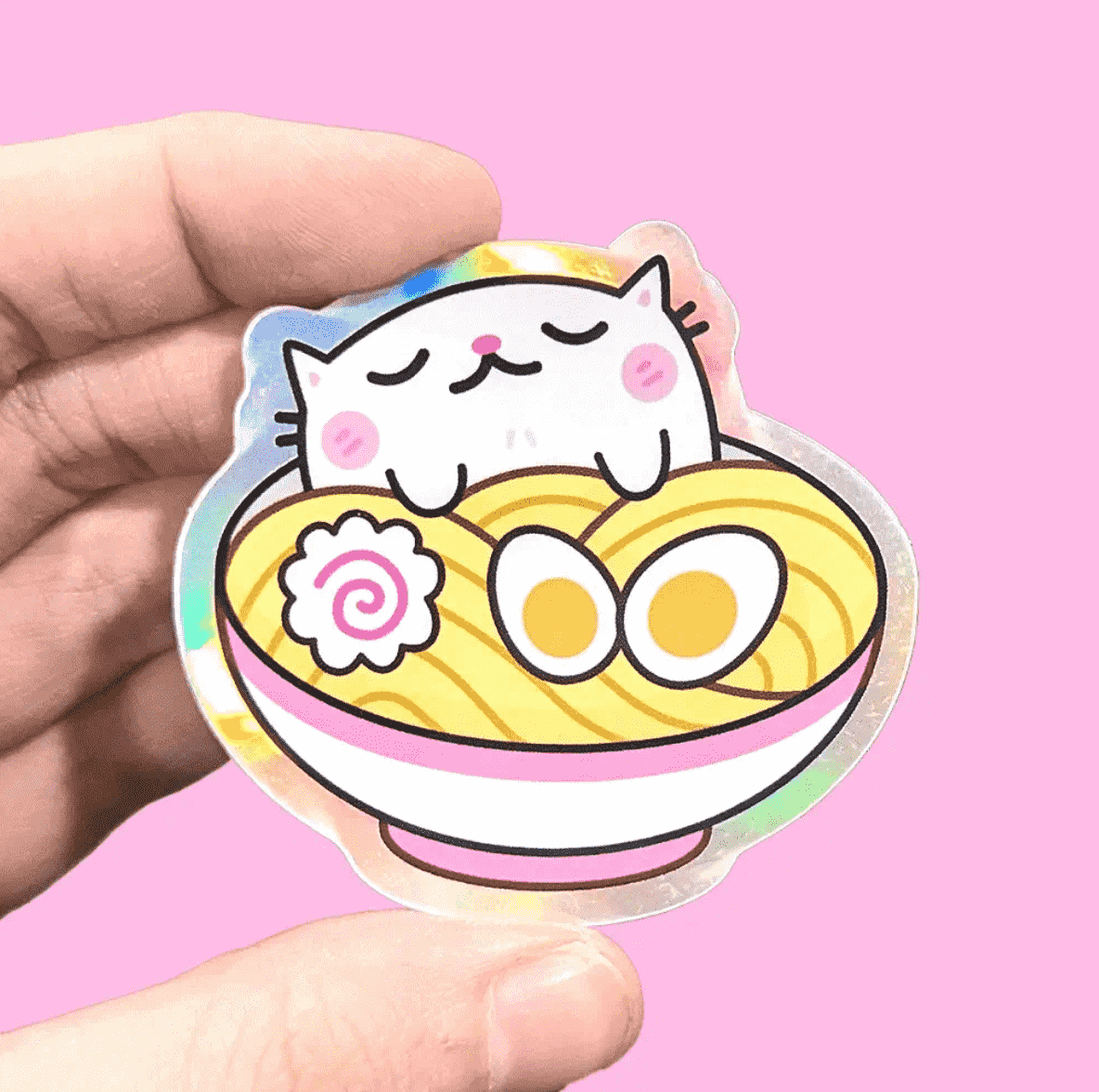 A holographic vinly sticker by Fox & Cactus of a small white cat with rosy cheeks holding a bowl of ramen. 