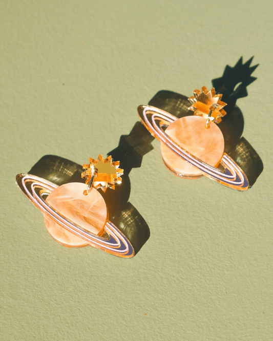 Flowature's Saturn Return Earrings. They have a gold stair at the top connecting to the air. A small gold hoop connects them to the saturn shape below featuring pink and gold. 