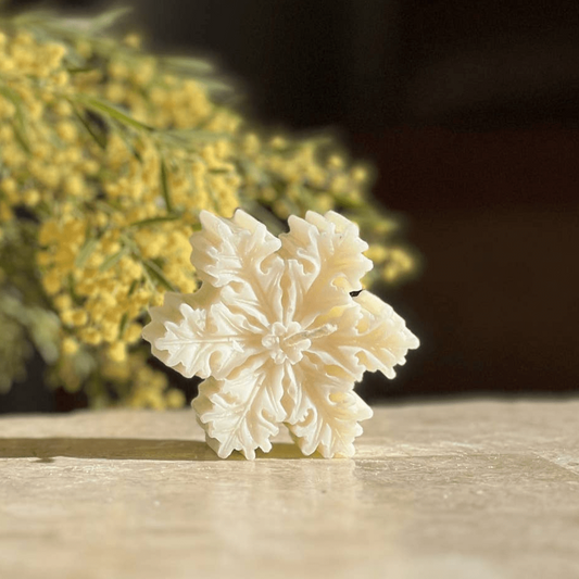 A beautiful handcrafted, pristine white snowflake candle with yellow wattle leaves in the foreground. 