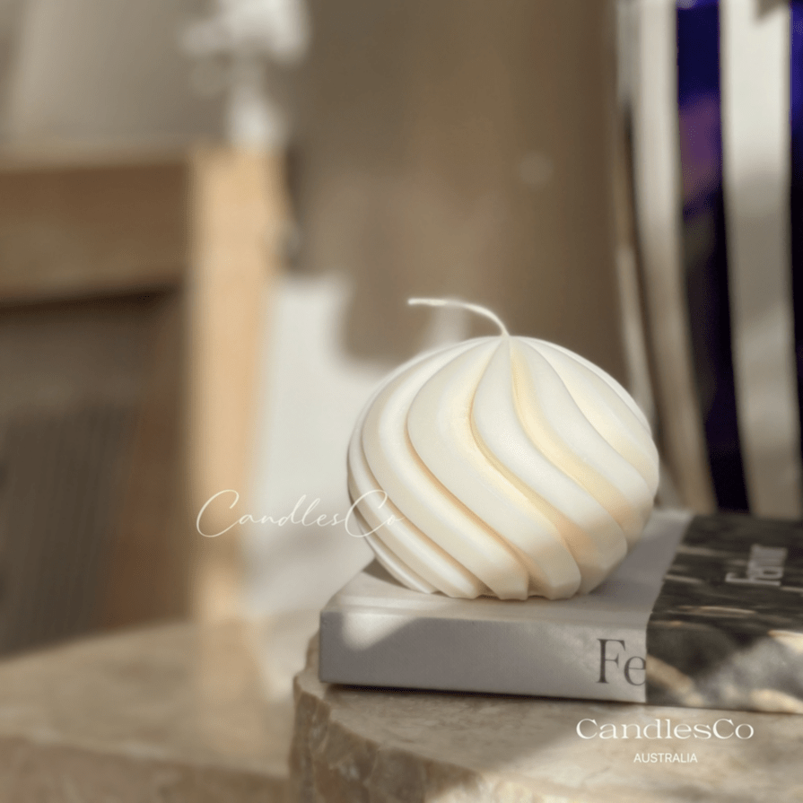 A photo of a cream coloured circular pillar candle in the shape of a spiral. It's resting on a book with a warm, cosy home in the background. 