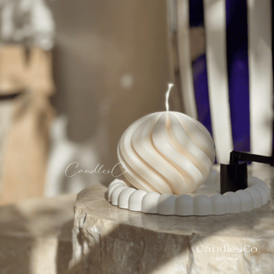 A photo of a cream coloured circular pillar candle in the shape of a spiral. It's resting on a book with a warm, cosy home in the background. 