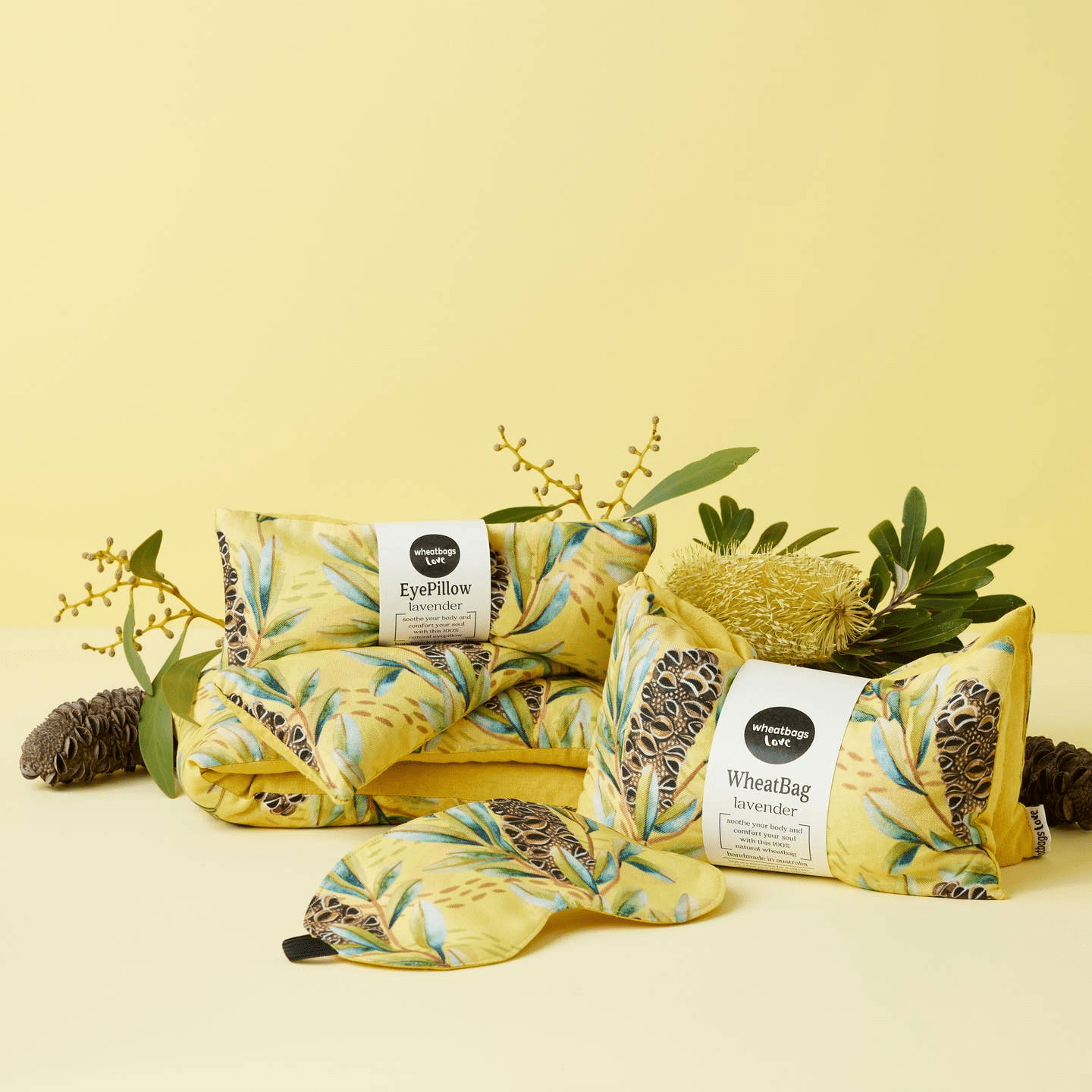 A collection of Wheatbag love's handmade eyepillows and wheatbags on a yellow backdrop with Australian fauna (including Eucalyptus leaves and Banksia pods in the foreground). The wheatbags themselves are yellow with green eucalyptus leaves and banksia pods on the print. 